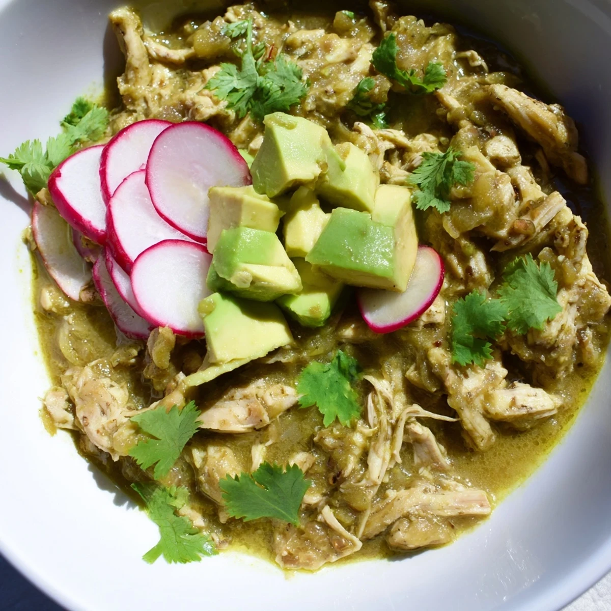 Homemade Chicken Chili Verde served in a rustic bowl, loaded with tender chicken, diced avocado, sliced radishes, and fresh cilantro for a festive touch.